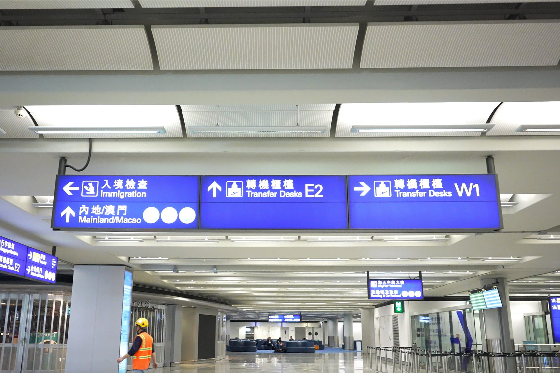 香港国際空港内の青い案内板。左は入境検査・中国本土/マカオ方面、中央は乗り継ぎカウンターE2、右は乗り継ぎカウンターW1への矢印が表示されている